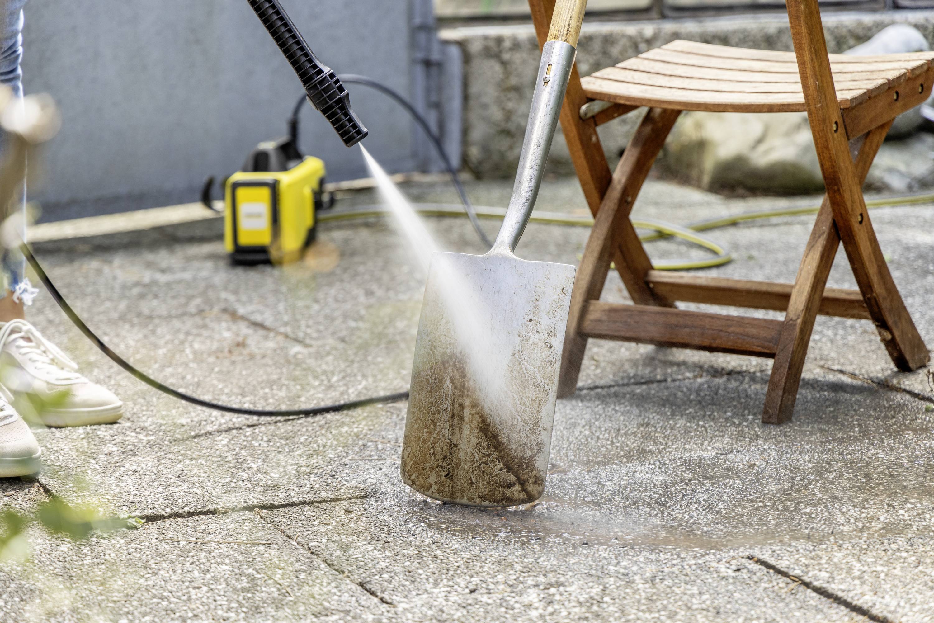 'Een persoon reinigt een schop met een hogedrukreiniger op een terras. Op de achtergrond staat een houten stoel.'