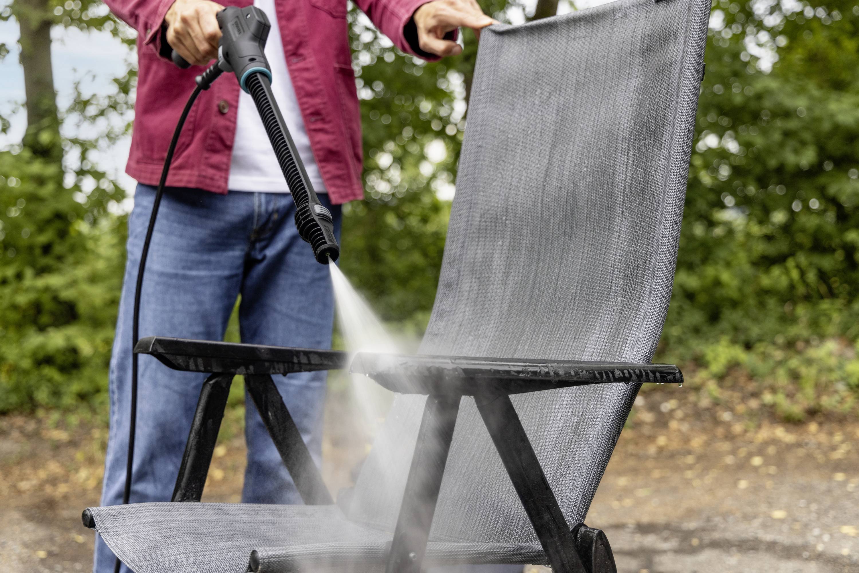 Een persoon reinigt een grijze tuinstoel met een hogedrukreiniger buiten, waarbij bomen op de achtergrond zichtbaar zijn.