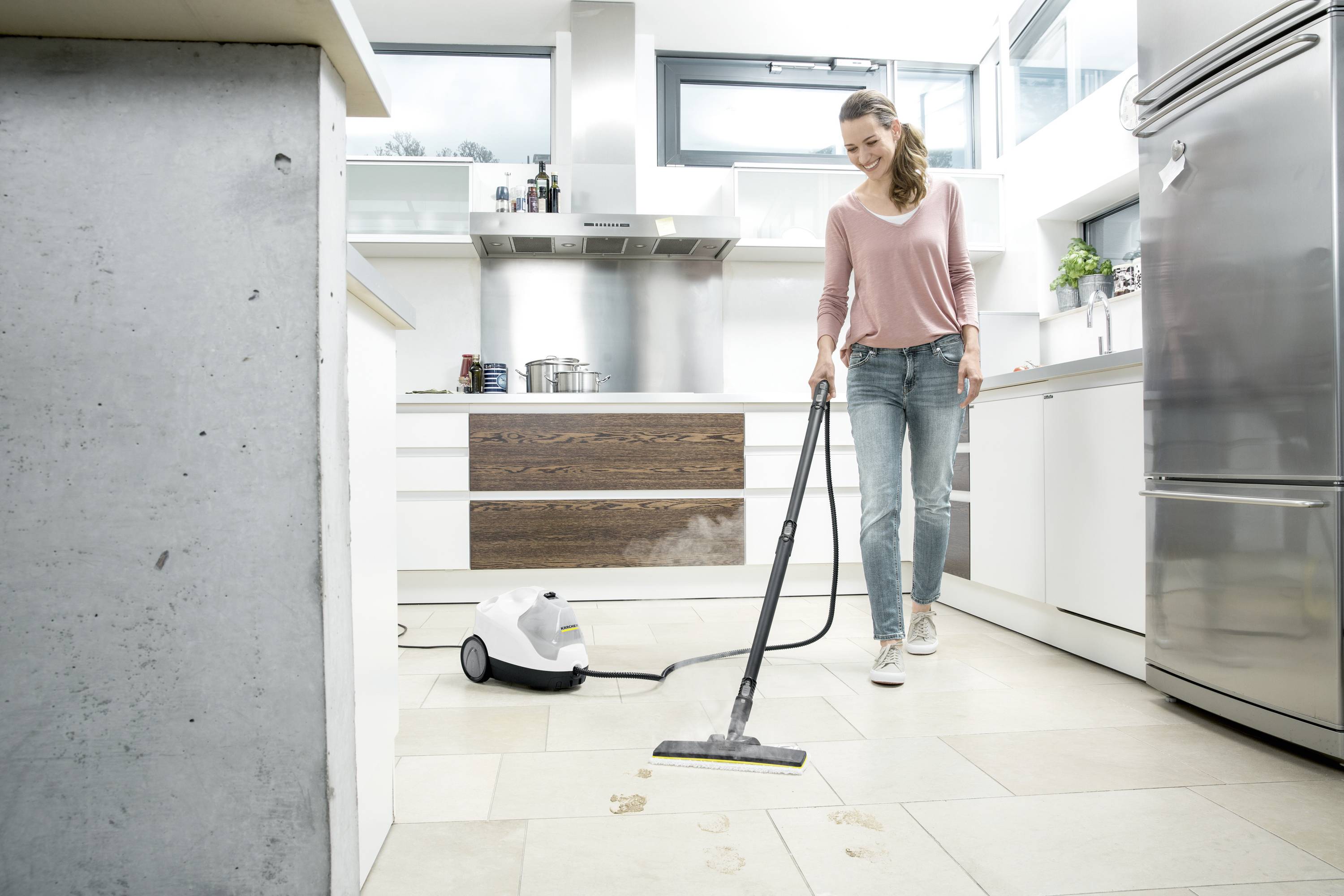 Een vrouw reinigt de keukenvloer met een stoomreiniger. De keuken is modern ingericht, met roestvrijstalen apparaten en veel licht.