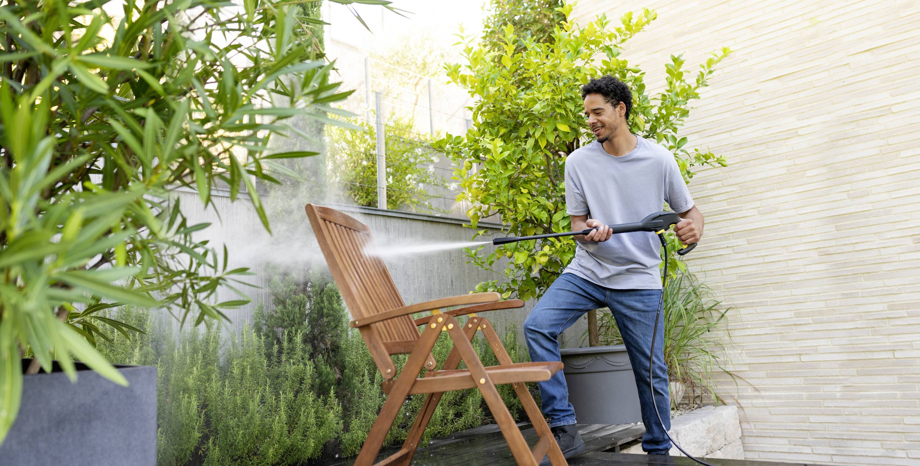 Een man reinigt een houten tuinstoel met een hogedrukreiniger op een terras naast groene planten.