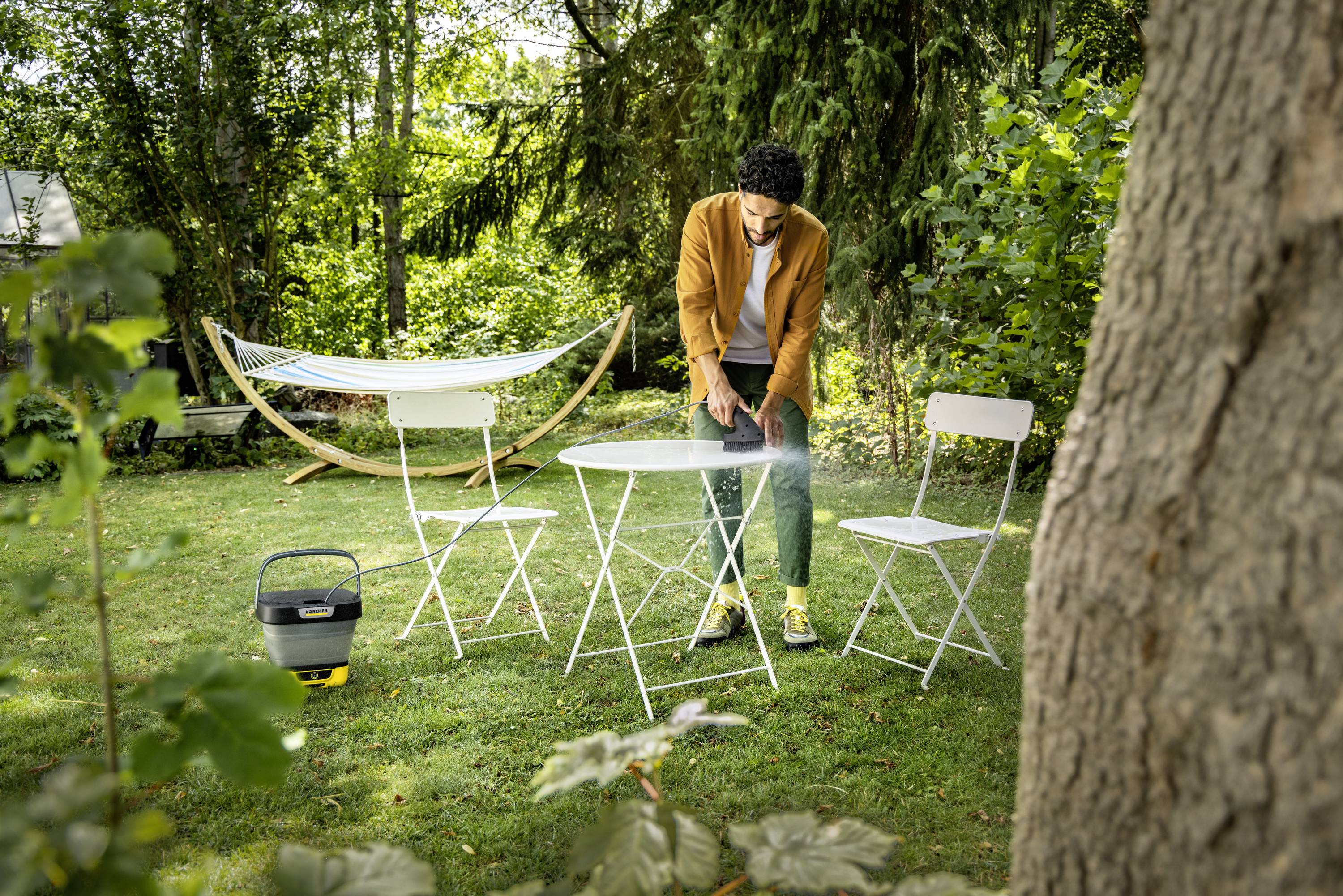 Een persoon steekt een barbecue aan op een tafel in een tuin, omringd door stoelen. Op de achtergrond is een hangmat zichtbaar.