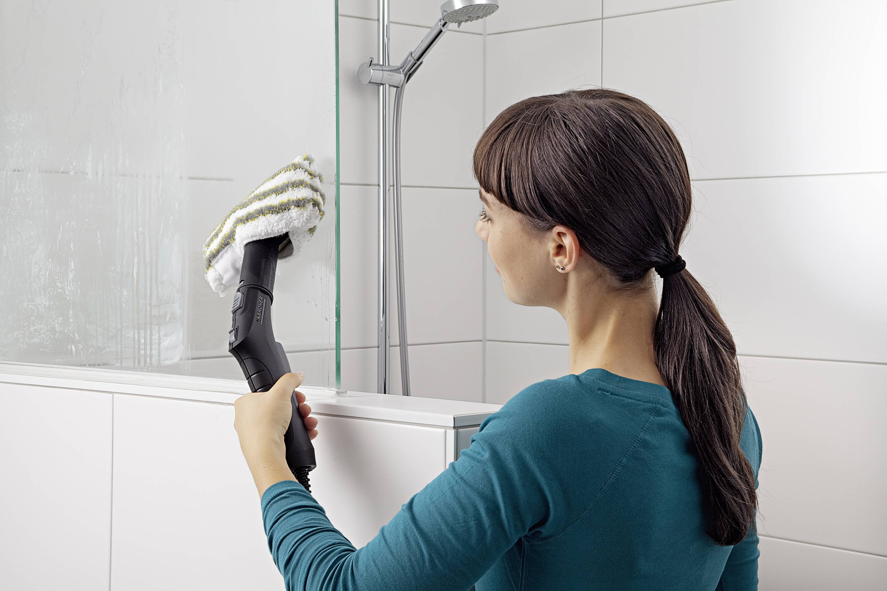 Een vrouw reinigt de glazen wand van een douchecabine met een stoomreiniger in de badkamer. Ze draagt een turquoise bovenkleding.