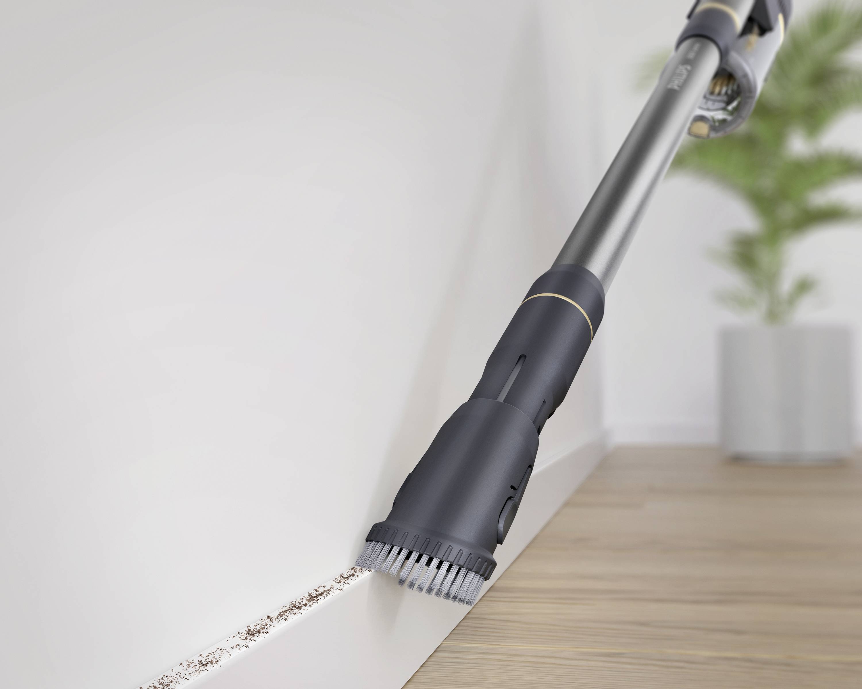 Een stofzuiger met borstelbijlage reinigt een plintstuk op een houten vloer in een ruimte met een plant op de achtergrond.
