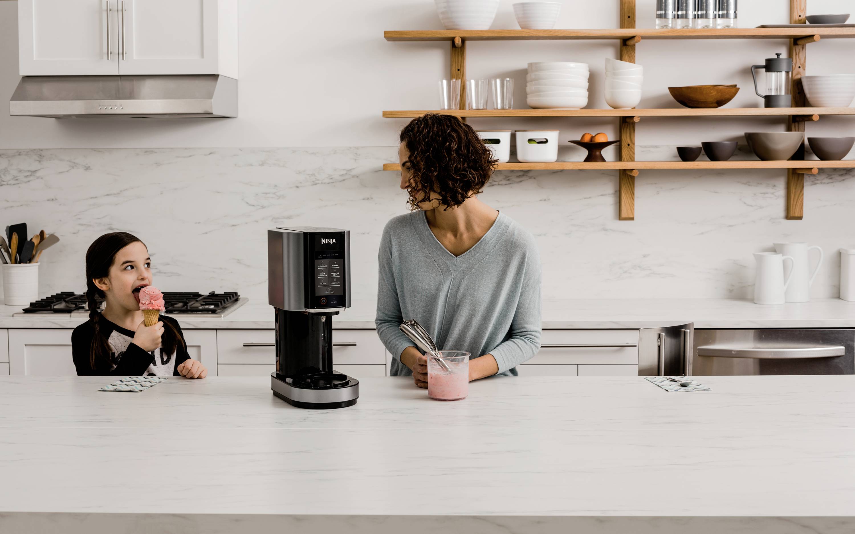 Een vrouw bereidt een drankje met een blender in de keuken, terwijl een meisje aan tafel zit en een ijsje eet.