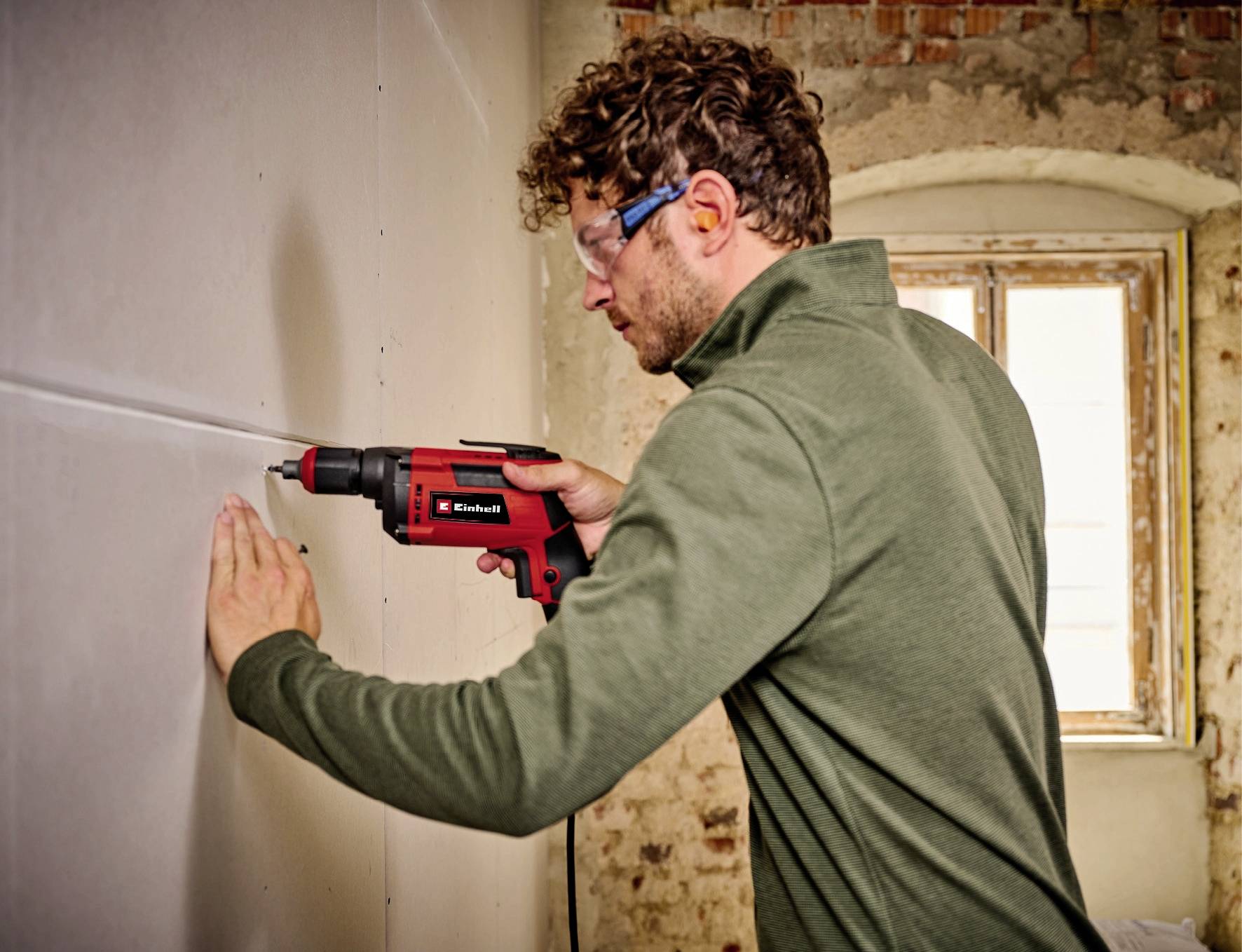 Een man boort in een muur in een ruimte die aan renovatie toe is. Hij draagt een veiligheidsbril en gebruikt een rood-zwarte boormachine.