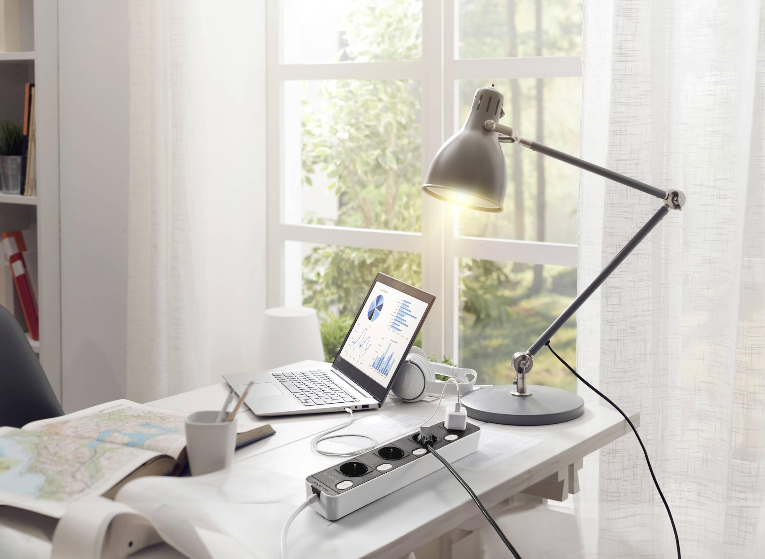 Een bureau bij het raam met laptop, lamp, stekkerdoos en een kaart. Zonlicht schijnt door witte gordijnen.