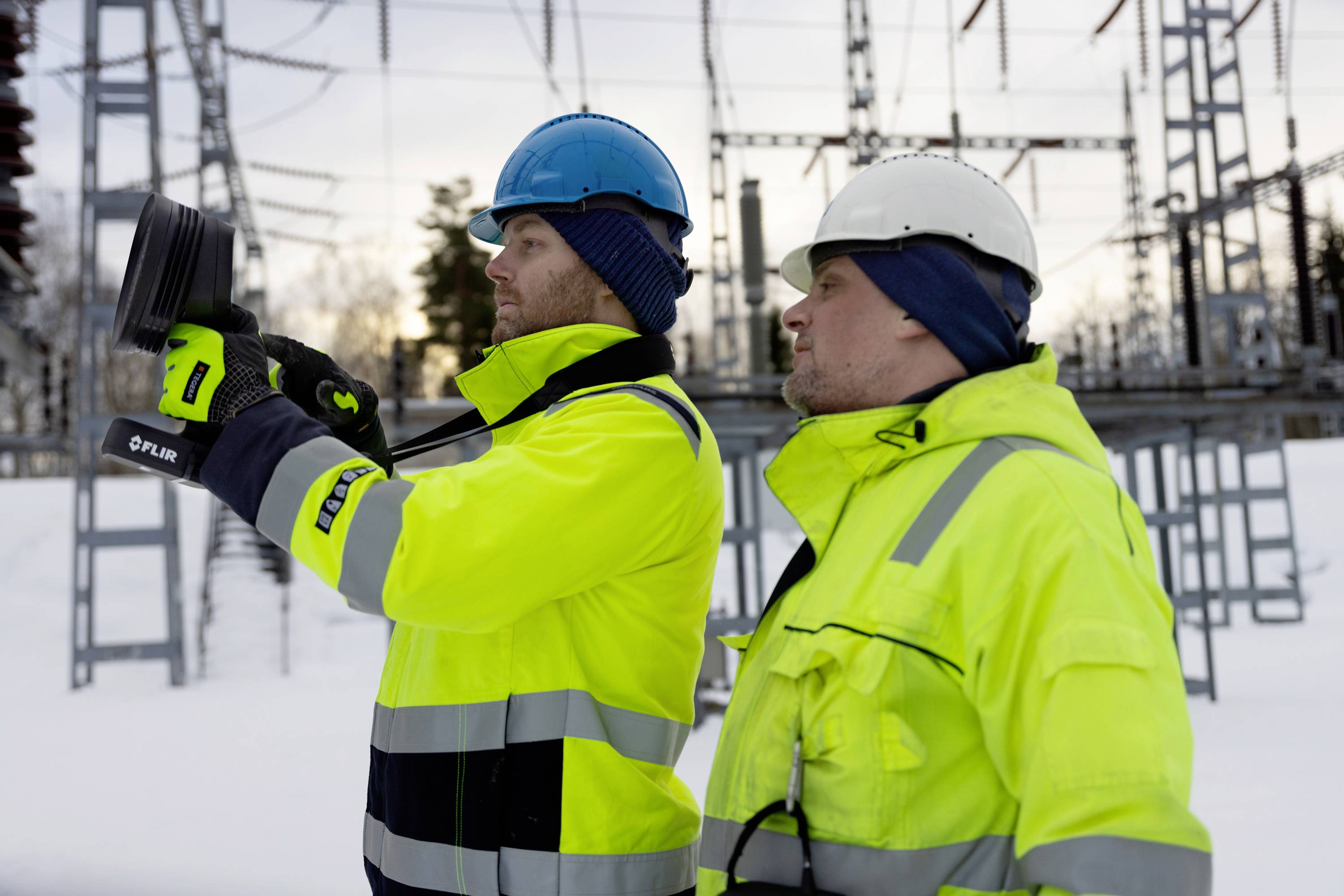 Twee personen in gele jassen en veiligheidshelmen inspecteren apparatuur in een met sneeuw bedekt elektriciteitsverdeelstation.