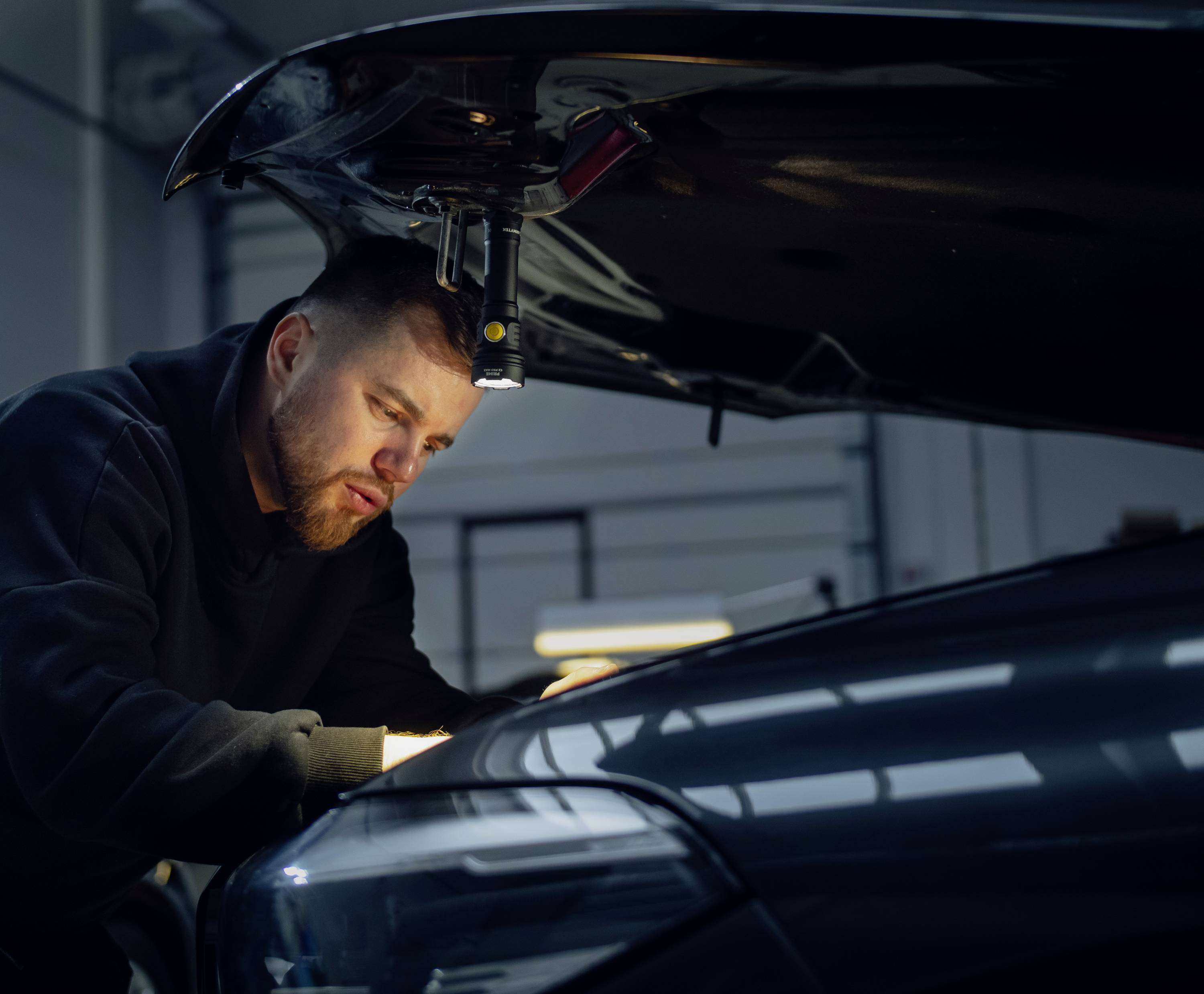 Een monteur werkt geconcentreerd onder de opgetilde motorkap van een auto in een werkplaats.
