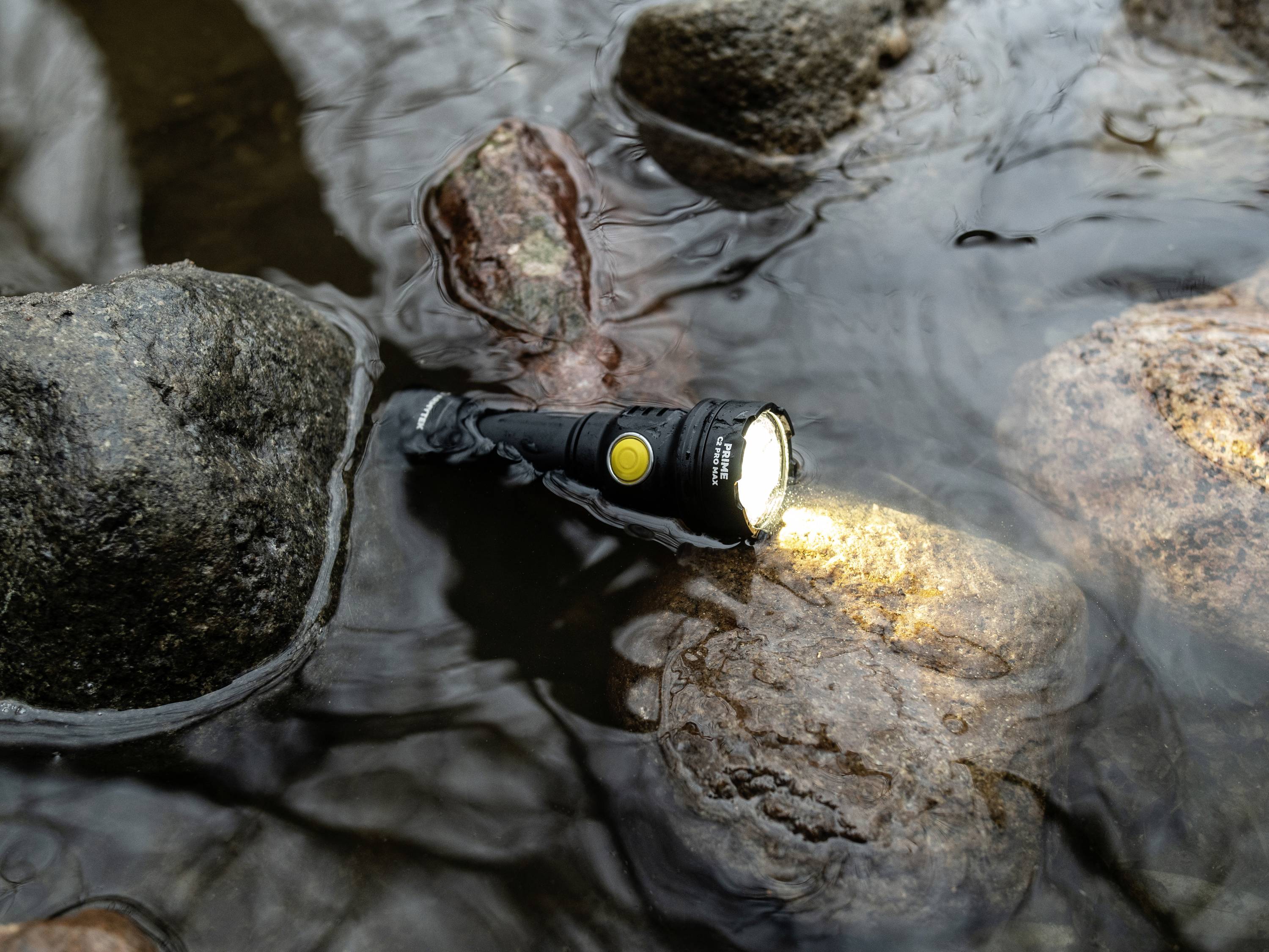 Een zaklamp ligt in een ondiep beekbedding tussen stenen. Hij schijnt in het water en werpt een lichtschijn op de stenen.