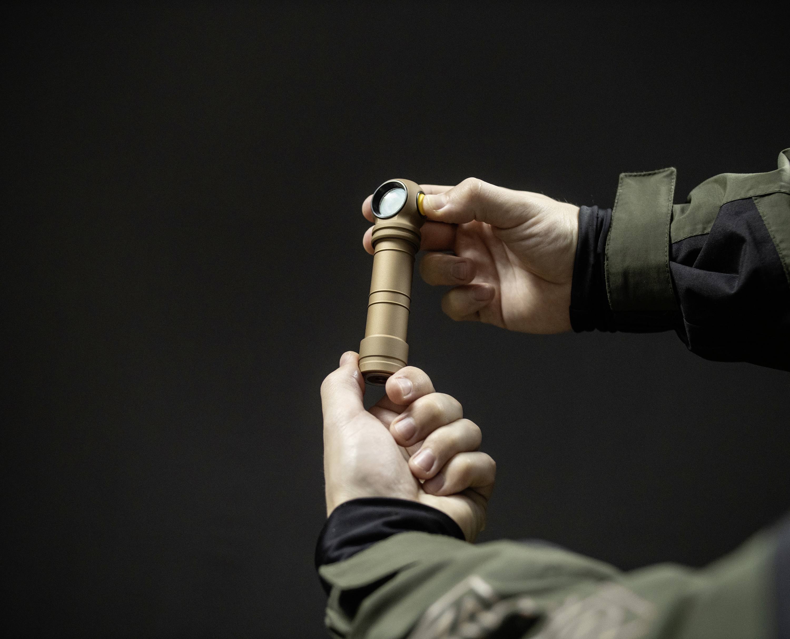 Een persoon houdt een kleine, bruine zaklamp in de hand, klaar voor gebruik. Op de achtergrond is een donkere, neutrale ruimte.