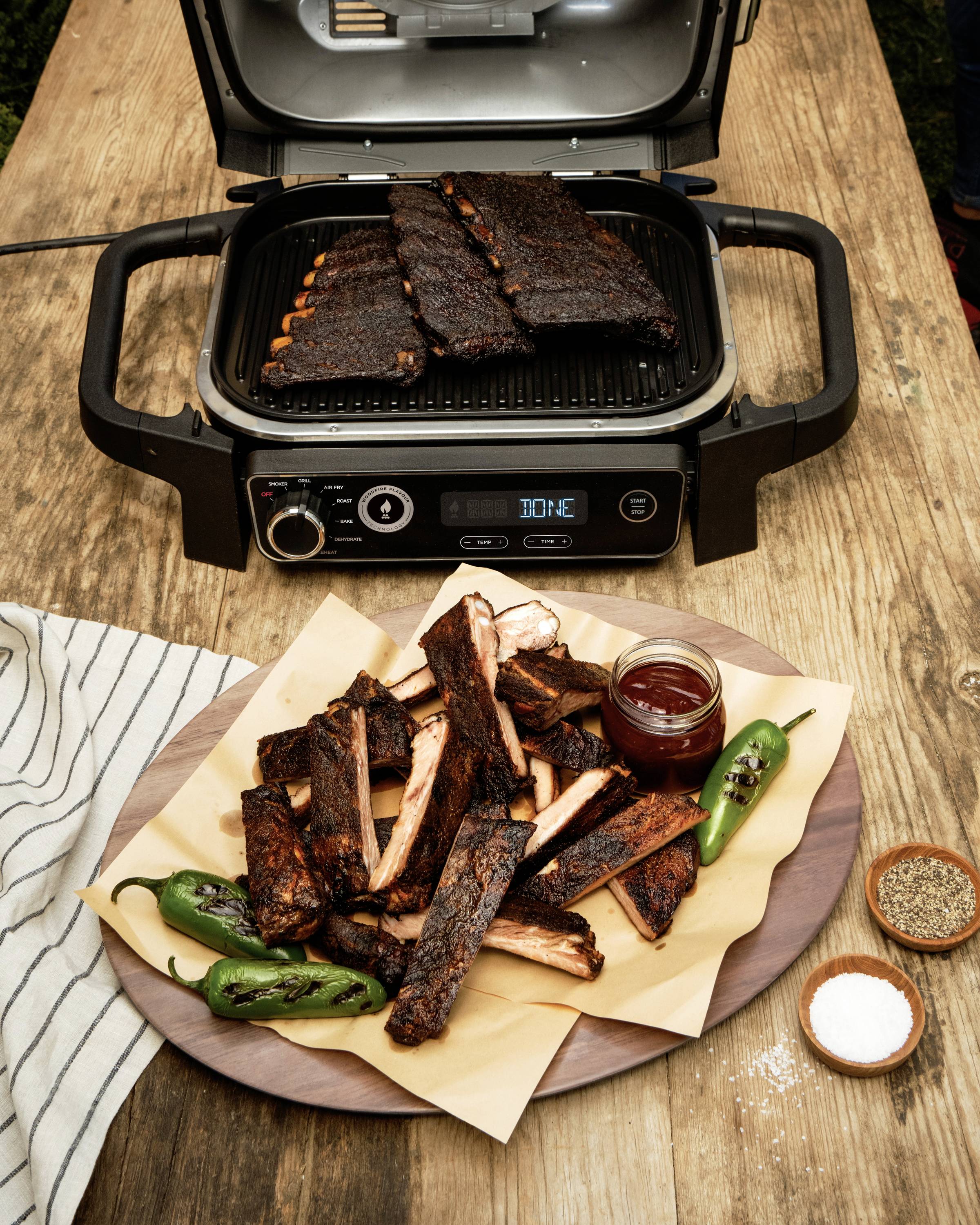 Een tafelgrill met gegrilde ribbetjes. Op een bord ernaast liggen afgewerkte ribbetjes, groene chilipepers en een kleine schaal met saus.