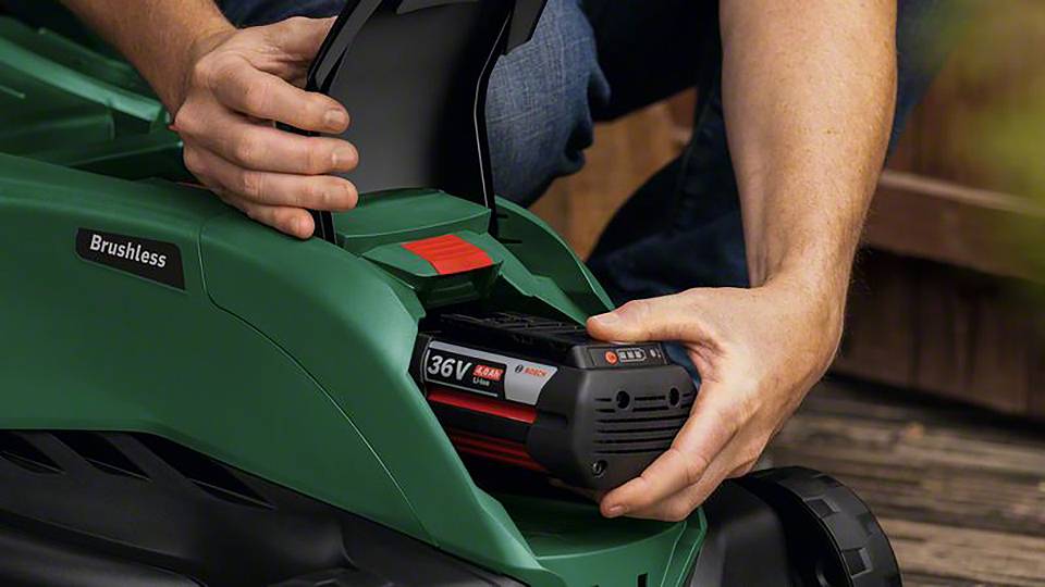 Een persoon plaatst een 36V accu in een groene, borstelloze grasmaaier. Handen houden de accu vast voor een houten achtergrond.