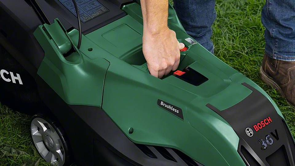 Een persoon steekt een batterij in een groene grasmaaier die op een grasveld staat. Het apparaat is gemarkeerd met 'Bosch'.