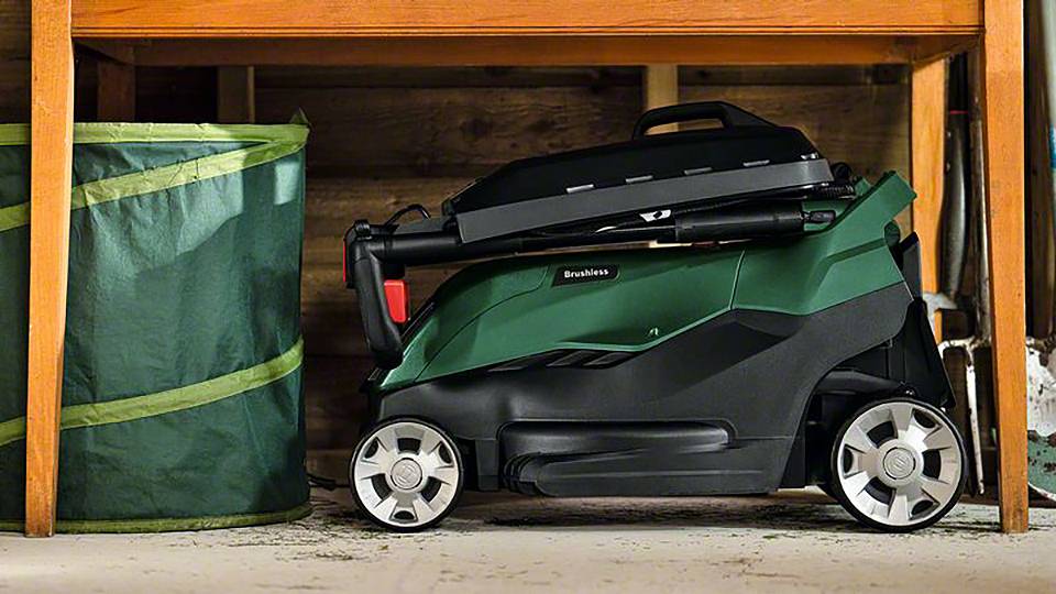 Een ingeklapte, groene grasmaaier onder een werkbank in een garage naast een grote, groene container.