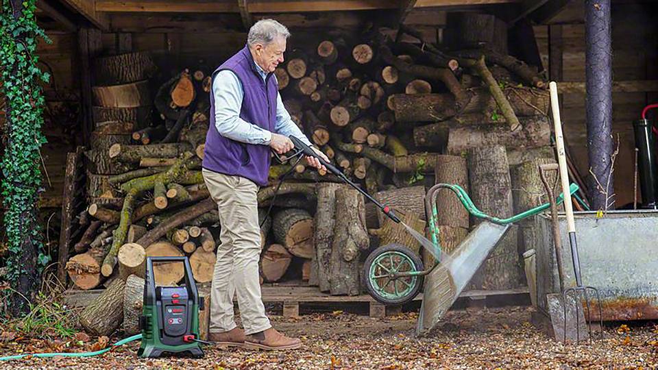 Een oudere man reinigt een kruiwagen met een hogedrukreiniger. Op de achtergrond zijn gestapelde brandhoutblokken te zien.