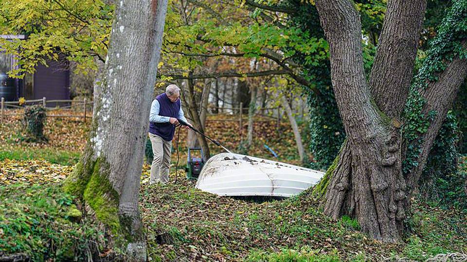 Oudere man in het park harkt herfstbladeren met een bladhark; omringd door grote bomen, bladeren in herfstkleuren verspreid over de grond.