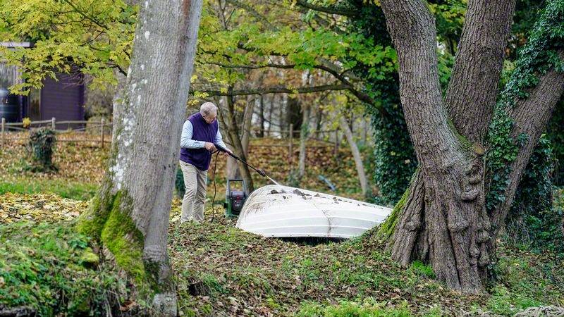 Een persoon harkt bladeren rond een omgekeerd roeiboot in een herfstelijke tuin, omgeven door kleurrijke bomen en grasvelden.