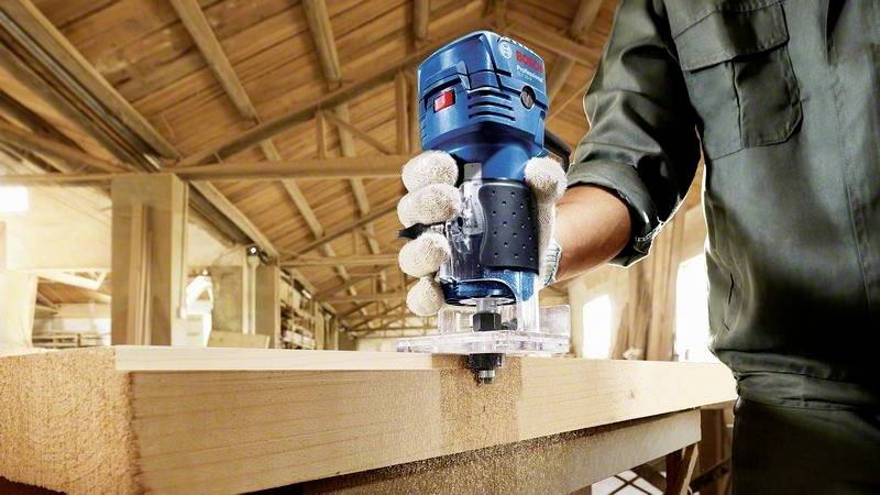 Een persoon in werkkleding gebruikt een blauwe bovenfrees om een houten plank nauwkeurig te bewerken in een werkplaats.