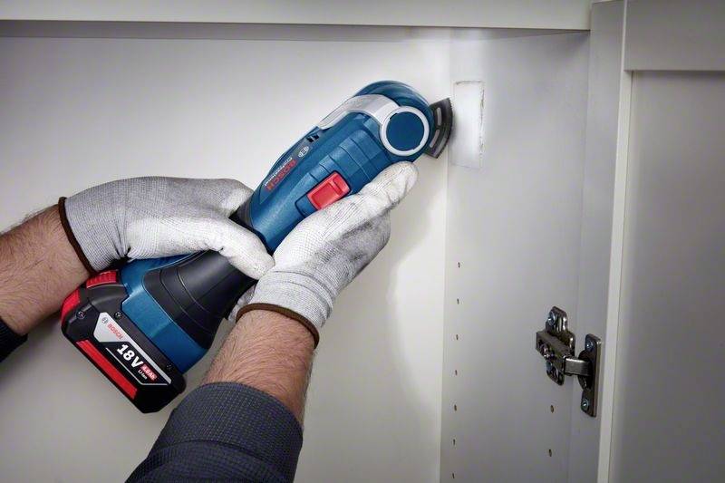 Een persoon houdt een blauw accusnijgereedschap vast en bewerkt de rand van een kast. Draagt handschoenen voor veiligheid.