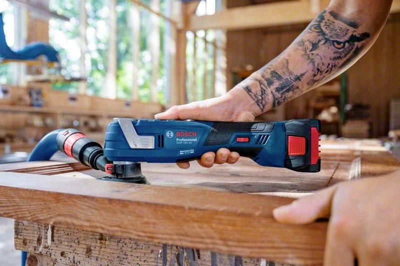 Een getatoeëerde persoon gebruikt een blauw multifunctioneel gereedschap van Bosch om een stuk hout te slijpen in een werkplaatsomgeving.