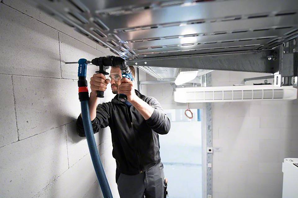 Een man installeert blauwe kabels in een technische ruimte. Hij staat op een ladder en werkt dicht bij het plafond.