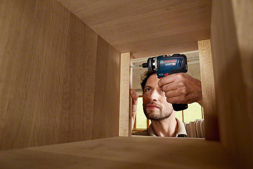 Een man monteert een houten rek. Hij gebruikt een blauwe accuschroevendraaier en werkt geconcentreerd.