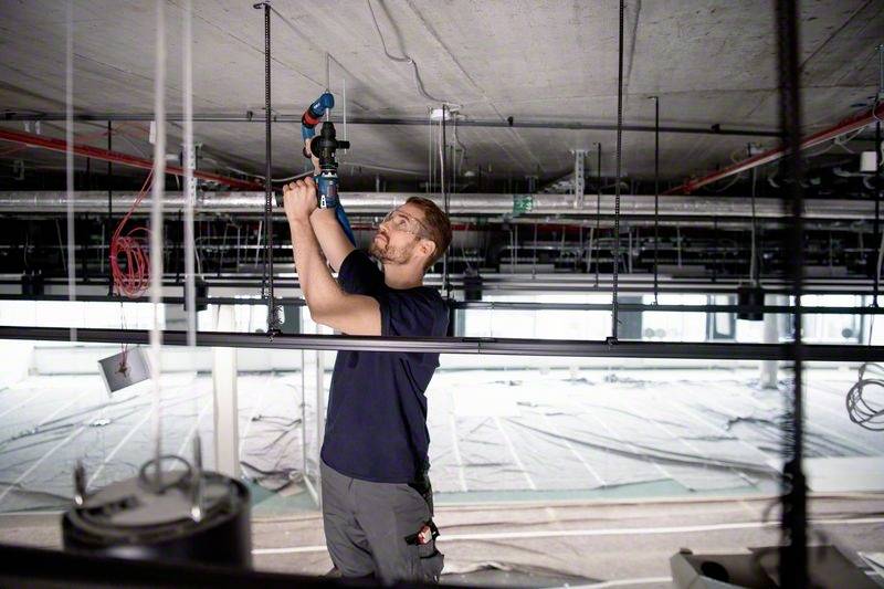 Een arbeider installeert leidingen aan het plafond in een grote, lege bouwplaats. Hij gebruikt een blauw en rood gereedschap.