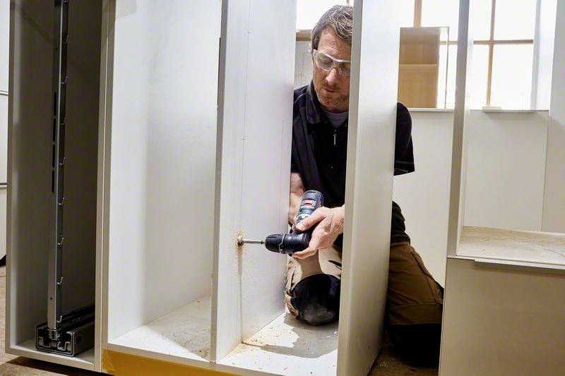 Een man gebruikt een boormachine om een houten meubelstuk in elkaar te zetten. Hij draagt een veiligheidsbril en knielt op de grond.