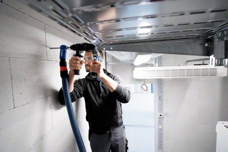 Een persoon installeert een blauwe ventilatiebuis in een technisch uitziende ruimte. Ze gebruikt gereedschap om de buis te bevestigen.