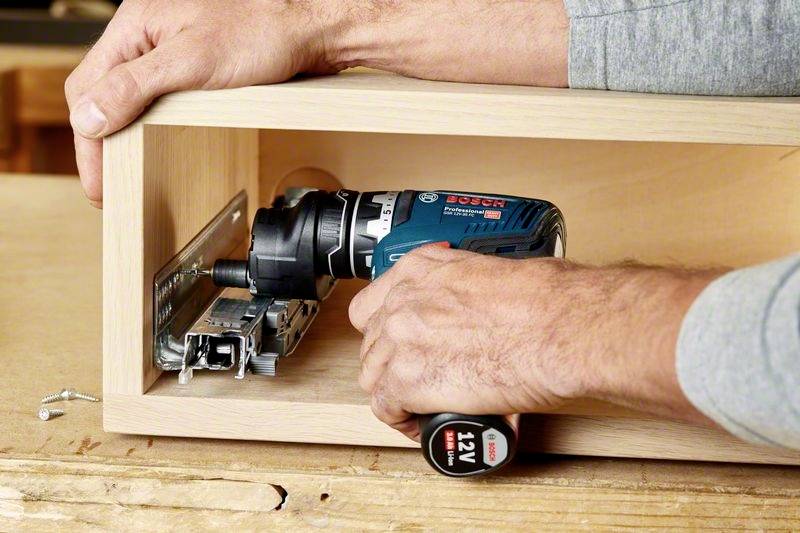 Een persoon monteert een metalen scharnier in een houten kist met een elektrische schroevendraaier.