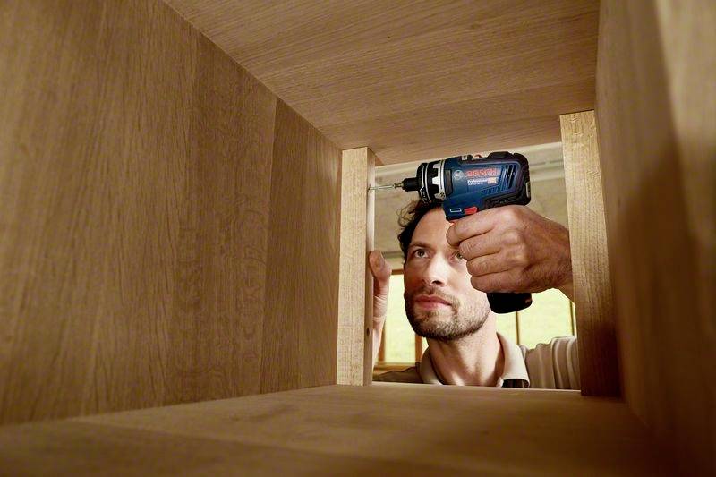 Een man werkt met een elektrische schroevendraaier aan een houten meubelstuk. Hij bevestigt onderdelen aan de binnenkant van het meubel.
