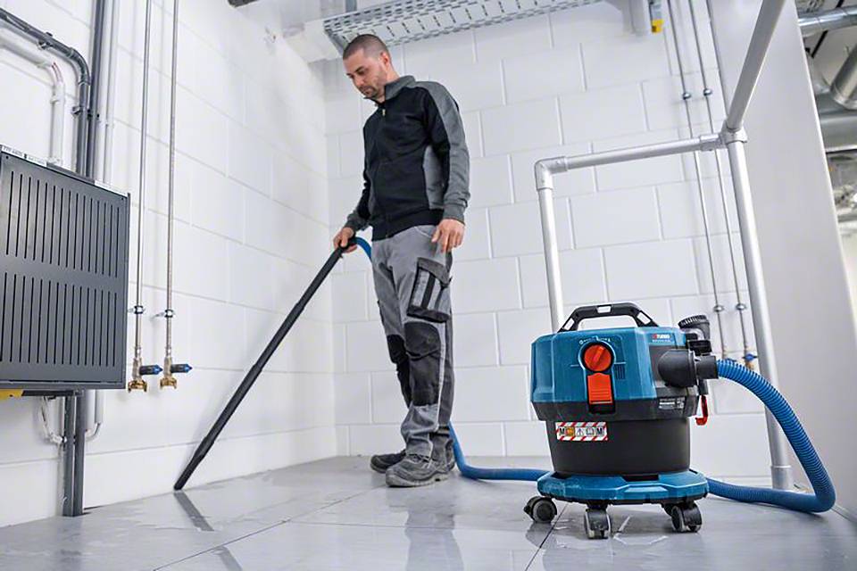 Een man staat in een kelder en reinigt de vloer met een nat- en droogzuiger. De ruimte heeft witte muren en verschillende leidingen.
