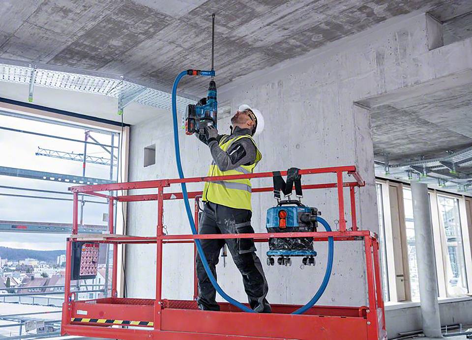 Een arbeider boort met een machine een gat in het plafond van een gebouw, staat op een hoogwerker, draagt beschermende kleding en een helm.