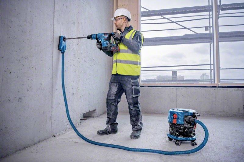 Een persoon in werkkleding boort in een betonnen muur op een bouwplaats. Een stofzuiger is aangesloten op de boormachine.