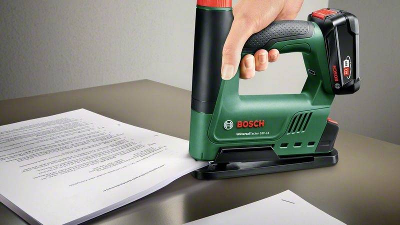 Een hand houdt een groene Bosch elektrische nietmachine, die gericht is op een stapel papier op een tafel.
