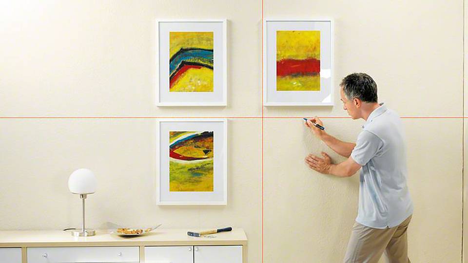 Een persoon hangt drie abstracte schilderijen aan een beige muur. Op de kast staan een lamp en schaaltjes.