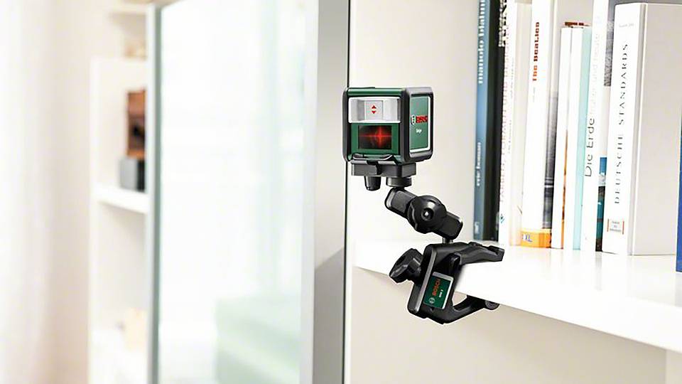 Een lasertelemetrie-apparaat is bevestigd aan een rek, projecteert een rood kruisdraad op de tegenoverliggende muur. Boeken op de achtergrond.