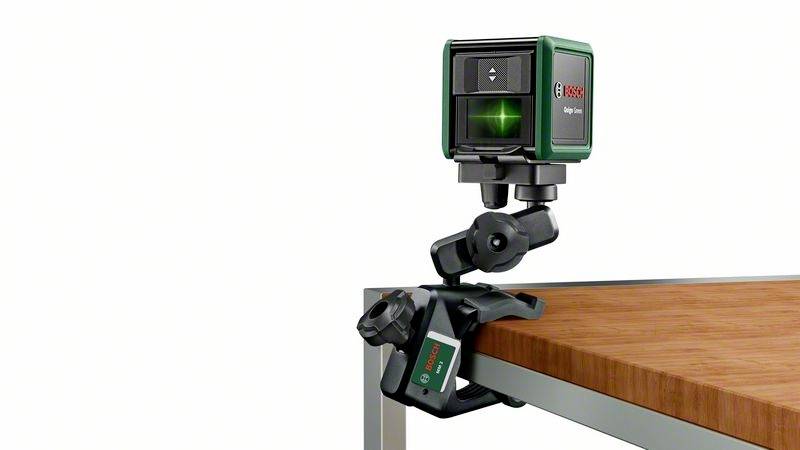 Een groene laserwaterpas is met een klem bevestigd aan de rand van een tafel. Het apparaat projecteert een groen kruis op de muur.
