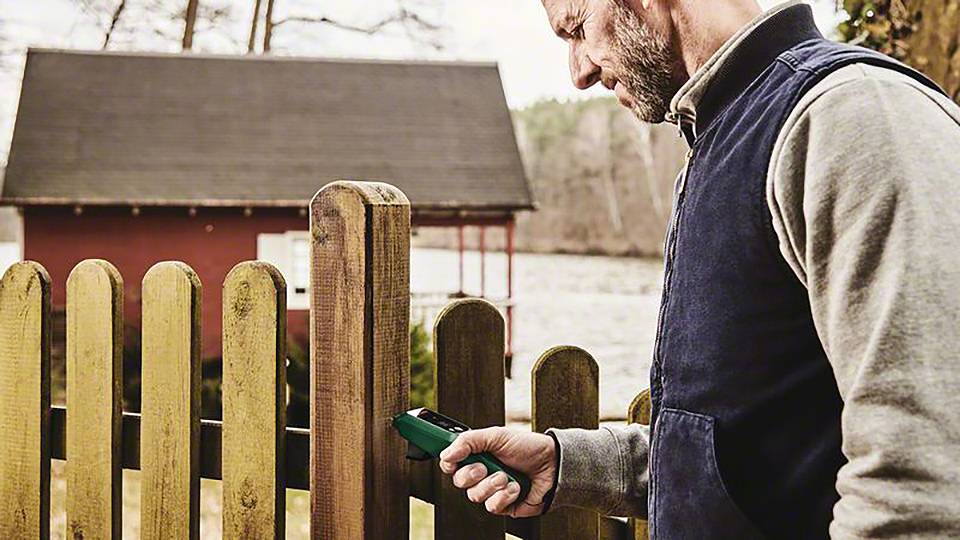 Een man meet de vochtigheid van een houten hek met een elektronisch apparaat. Op de achtergrond is een meer en een rood huis.
