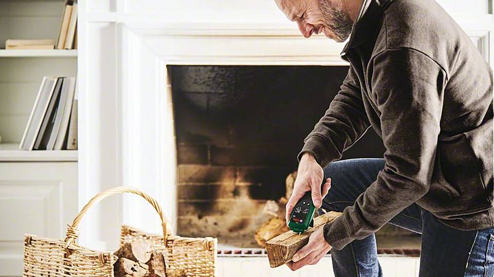 Een man meet een stuk brandhout met een digitaal apparaat in een woonkamer. Op de achtergrond flikkert een haardvuur.