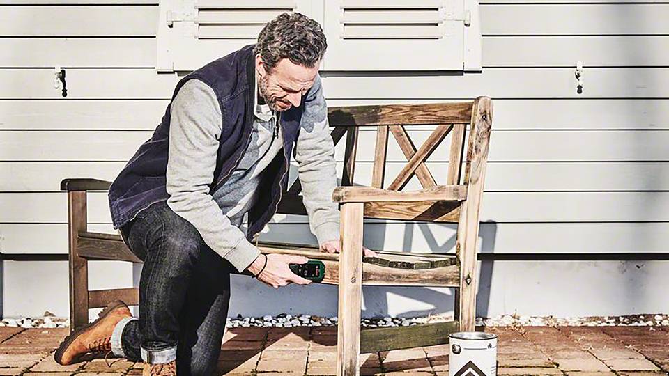 Een man repareert buiten een houten bank met een gereedschap, voor een licht houten huis. Naast hem staat een verfblik.