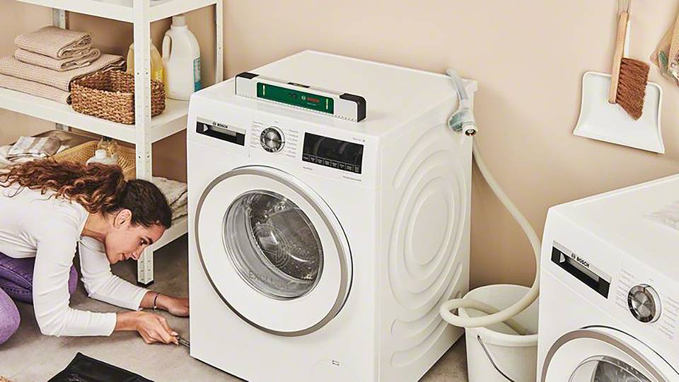 Een vrouw knielt naast een wasmachine en reinigt de vloer. Op de achtergrond zijn rekken met handdoeken en schoonmaakmiddelen te zien.