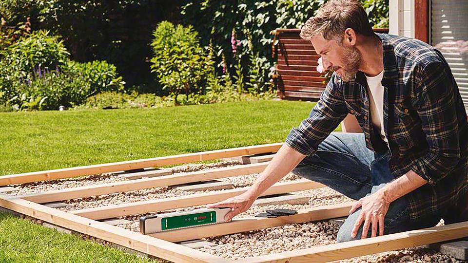 Een man knielt in de tuin en controleert met een waterpas het frame van houten balken dat op grind in het gazon ligt.