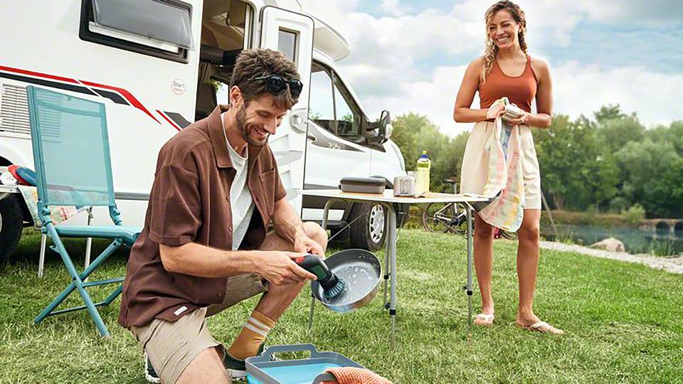 Een man doet de afwas bij een caravan terwijl een vrouw naast hem staat te lachen. Op de achtergrond zijn bomen en een rivier te zien.