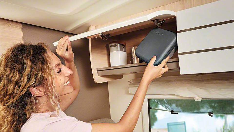 Een vrouw bergt een donker, rechthoekig etui op in een kastje van een camper. Ze kijkt glimlachend naar het etui.