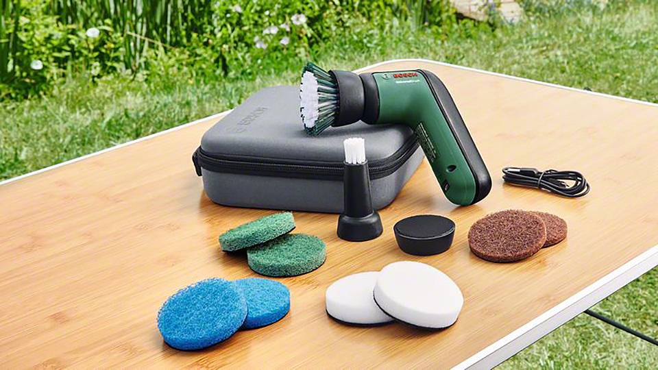 Een groen elektrisch reinigingsapparaat met verschillende opzetstukken en een kabel ligt op een houten tafel buiten voor een gazon.