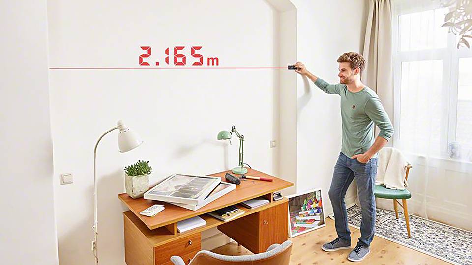 Een man meet de breedte van een kamer met een digitaal afstandsmeetapparaat. De meting toont 2,165 meter aan.