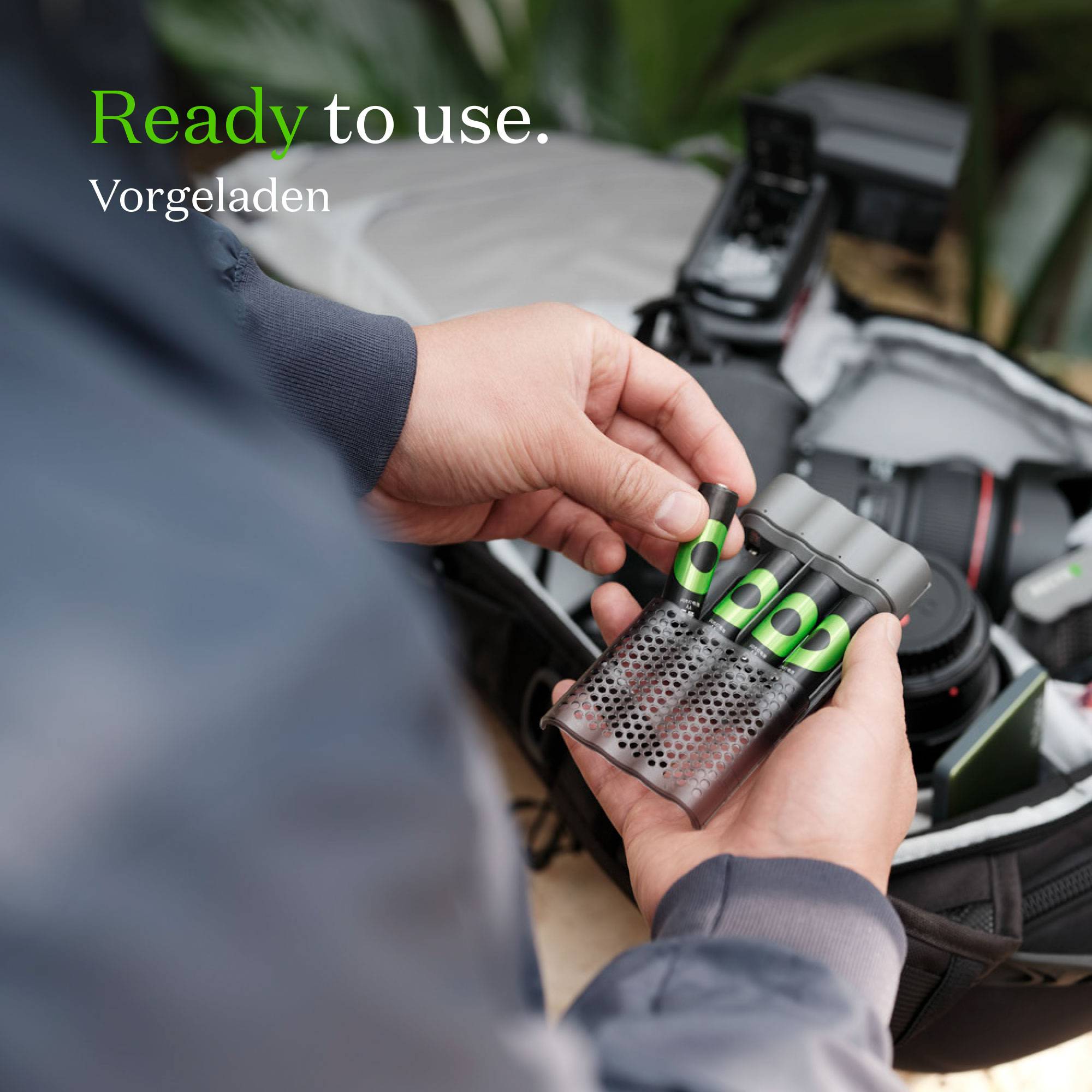 Een persoon houdt een open batterijvak met vier klaargelegde batterijen. Op de achtergrond is fotoapparatuur zichtbaar. Tekst in beeld: 'Klaar voor gebruik. Voorgeladen'.