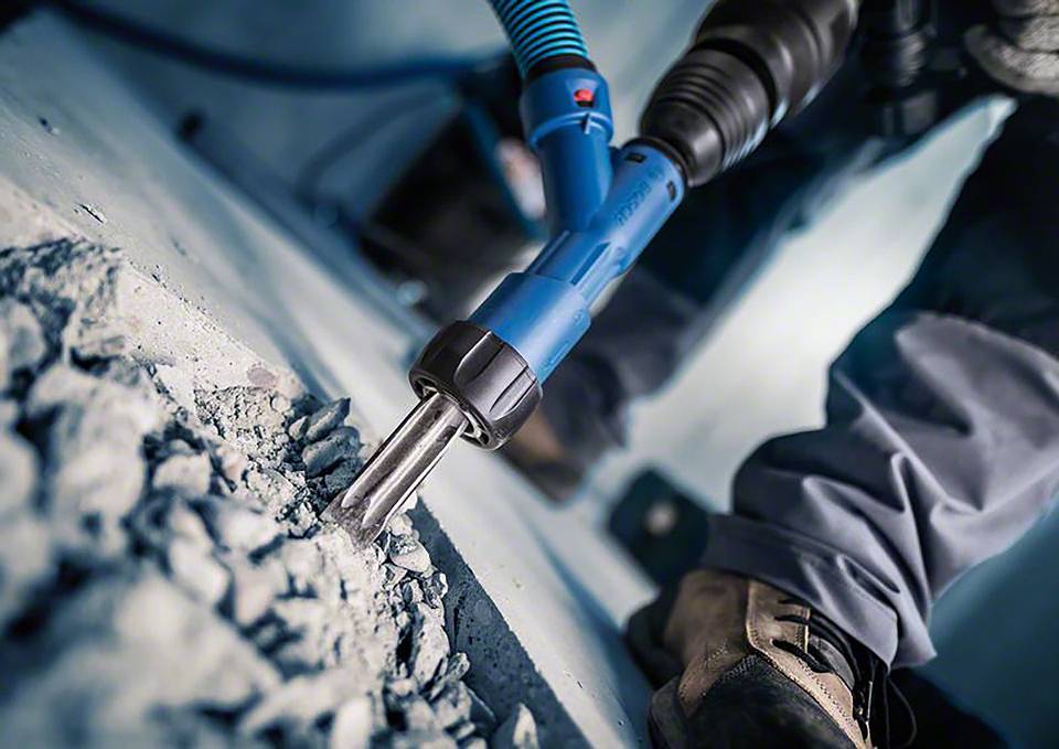 Een persoon gebruikt een blauwe pneumatische hamer om beton te breken. Close-up van gereedschap en schoenen, geen gezichten zichtbaar.