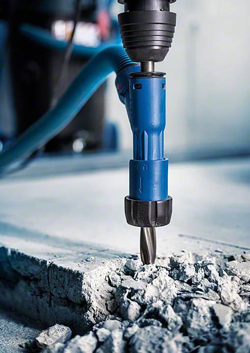 Een blauwe boorhamer breekt beton kapot. De focus ligt op de boorpunt, die betonbrokken vergruizelt. Bouwplaatsomgeving.